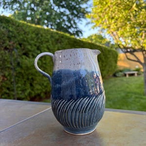 May include: A ceramic pitcher featuring a two-tone glaze with a speckled cream top and a deep blue bottom. The lower section of the pitcher displays a textured pattern of carved vertical grooves, and it includes a curved handle and a small spout.