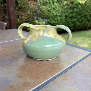 May include: A handcrafted ceramic vase with two loop handles. The vase has a rounded body and is glazed in shades of green and yellow. The handles have a textured, rope-like appearance. The vase is on a brown tiled surface.