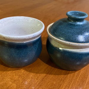 May include: A two-piece ceramic serving set. The set includes a creamer with a spout and a lidded container. Both pieces are blue with a speckled white rim and a white interior. The set is likely for serving condiments or sauces.