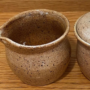 May include: A speckled brown ceramic creamer with a spout and a matching sugar bowl with a lid. The pottery has a rustic, handmade appearance. The creamer and sugar bowl are set on a wooden surface.