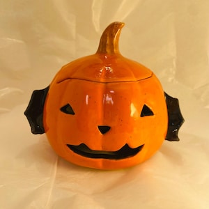 Peut inclure: Une lanterne en céramique en forme de citrouille, peinte en orange brillant avec des accents noirs. La citrouille a un visage sculpté avec des yeux triangulaires et une bouche incurvée. Des poignées noires en forme de chauve-souris sont de chaque côté.