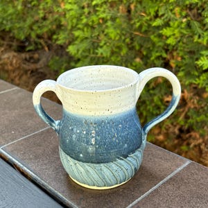 May include: A ceramic mug featuring two handles and a two-tone glaze. The upper portion is a speckled off-white, transitioning into a deep blue midsection and a light blue base with a textured, swirling carved pattern around the bottom.