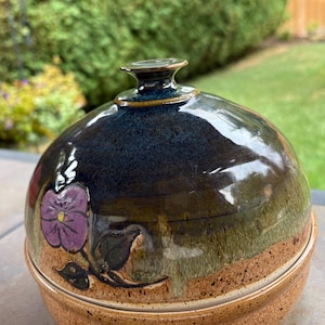May include: A handmade ceramic lidded bowl with a dark blue and brown glaze. The bowl features a purple flower design and a textured, speckled base. The lid has a small handle. This decorative bowl is suitable for serving or display.