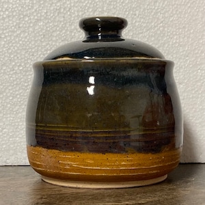 May include: A handmade ceramic jar with a lid. The jar has a dark brown glaze at the top, a lighter brown band in the middle, and a tan base. The lid is topped with a small knob for easy handling.