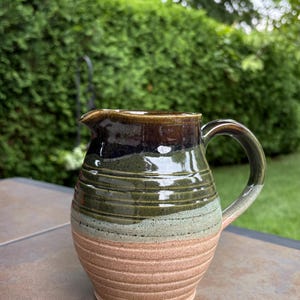 May include: A handcrafted ceramic jug with a dark brown rim, fading into green and tan bands. It features a curved handle and a pouring spout. The textured surface enhances its rustic appeal. Green foliage forms the background.