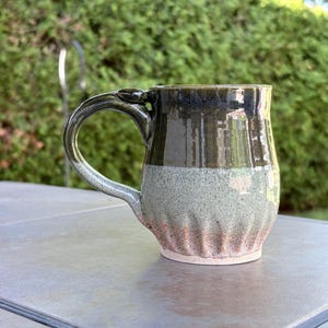 May include: A ceramic mug featuring a glossy dark green upper section that transitions into a lighter sage green middle, ending in a textured, fluted base with a warm terracotta hue. The mug includes a curved handle and a smooth, glazed finish.