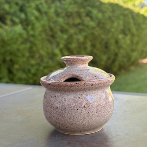 Peut inclure: Un petit pot en céramique rond avec un couvercle assorti, présentant un émail beige et brun moucheté. Le pot a un corps large et incurvé et un petit bouton sur le couvercle, qui comprend une encoche pour laisser reposer une cuillère.