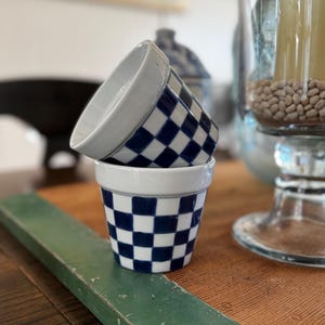 May include: Two small ceramic flower pots stacked on a wooden surface. The pots have a white rim and a blue and white checkered pattern. The background includes a green wooden plank and a glass candle holder.