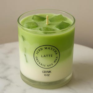 K&ouml;nnte beinhalten: Eine Sojawachskerze in einem klaren Glas, die einem eisgek&uuml;hlten Matcha Latte nachempfunden ist. Die Kerze hat einen geschichteten Effekt mit einer cremewei&szlig;en Basis, einer leuchtend gr&uuml;nen Mitte und gr&uuml;nen Wachsw&uuml;rfeln oben. Das Etikett lautet "Iced Matcha Latte, Organic Soy Wax, 10 oz".
