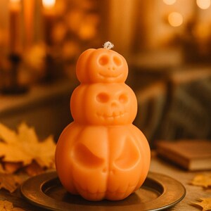 Può includere: Una candela arancione a forma di zucca impilata, con facce di jack-o'-lantern intagliate. La candela è su un piatto di bronzo, circondata da foglie autunnali. Lo sfondo presenta candele sfocate e illuminazione calda, per Halloween.