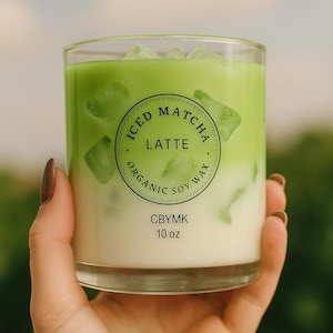 K&ouml;nnte beinhalten: Ein Glas Eiskaffee-Matcha-Latte mit Eisw&uuml;rfeln, gehalten von einer Person. Das Getr&auml;nk ist geschichtet mit einer gr&uuml;nen Oberseite und einer wei&szlig;en Unterseite. Das Glas tr&auml;gt die Aufschrift "Iced Matcha Latte, Organic Soy Wax, CBYMK, 295 ml."