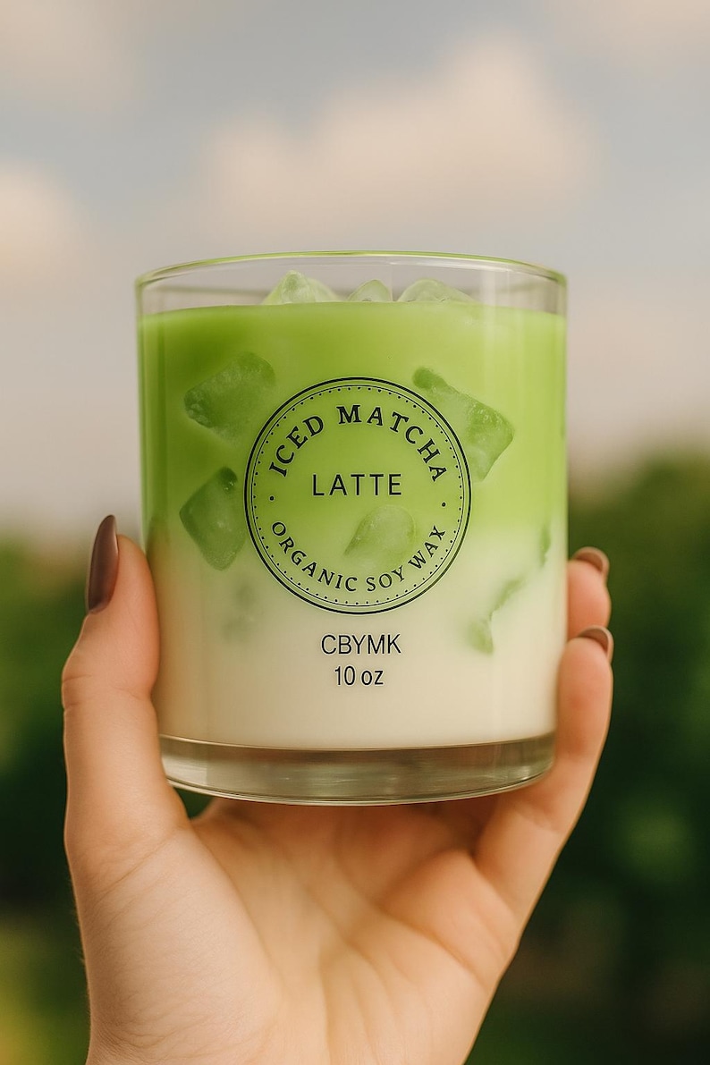 K&ouml;nnte beinhalten: Ein Glas Eiskaffee-Matcha-Latte mit Eisw&uuml;rfeln, gehalten von einer Person. Das Getr&auml;nk ist geschichtet mit einer gr&uuml;nen Oberseite und einer wei&szlig;en Unterseite. Das Glas tr&auml;gt die Aufschrift "Iced Matcha Latte, Organic Soy Wax, CBYMK, 295 ml."