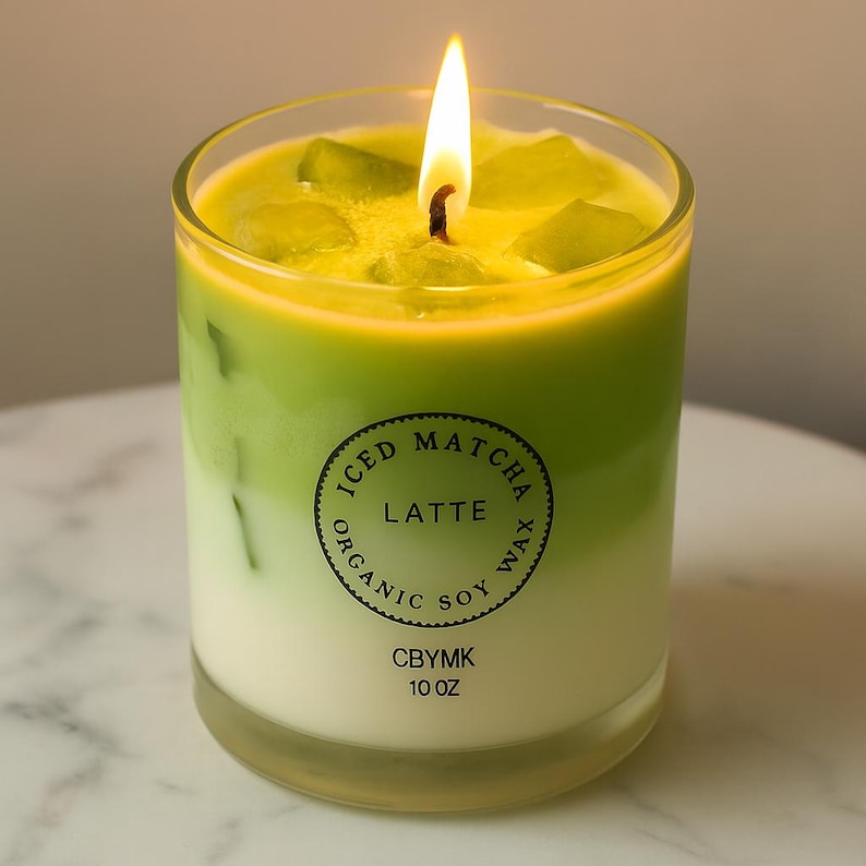 K&ouml;nnte beinhalten: Eine brennende Kerze in einem klaren Glas, die einem Eiskaffee-Matcha-Latte &auml;hnelt. Die Kerze hat einen Schichteffekt mit gr&uuml;nem und wei&szlig;em Wachs, und das Etikett lautet "Iced Matcha Latte Organic Soy Wax" und "10 oz".