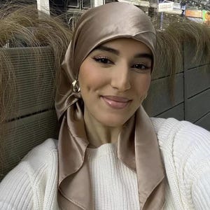 May include: A woman wearing a beige satin headscarf and gold hoop earrings. She is also wearing a white sweater. The headscarf is draped over her head and shoulders. The background includes outdoor elements.