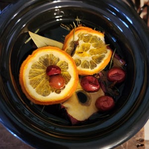 Puede incluir: Una olla de cocción lenta negra llena de rodajas de naranja secas, clavos y bayas rojas. Las rodajas de naranja están dispuestas en un círculo alrededor del centro de la olla.