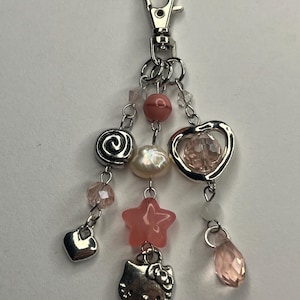 May include: A silver-toned metal keyring featuring three dangling strands attached to a lobster clasp. The strands include pink and clear faceted beads, a white pearl-like bead, a silver swirl charm, a silver heart, a pink star, and a cat charm.