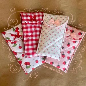 May include: Four quilted fabric cases in various patterns: cherries, red gingham, floral, and strawberries. Each case has a flap closure and a bow. The cases are arranged on a brown surface with decorative swirls and the text "Sweethearts Order".