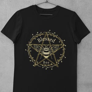 May include: Black t-shirt with a gold pentagram and bee design. The design includes the text "Blessed Bee".