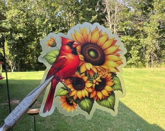 May include: A vibrant sticker featuring a red cardinal perched on a cluster of sunflowers. The sunflowers display yellow petals and dark brown centers, with green leaves. The sticker is held by a pair of pliers against a backdrop of green grass and trees.