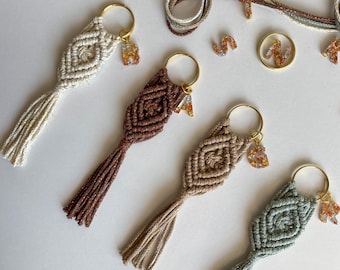 May include: Four handmade macrame keychains with tassels in beige, brown, light brown, and light blue. Each keychain has a gold ring and a small resin letter charm.