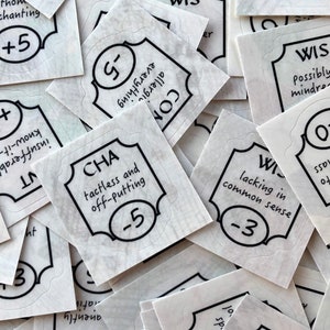 May include: A pile of white square stickers with black text and borders. The stickers have different words and numbers on them, such as "DEX", "WIS", "STR", "CON", "CHA", and "INT". Some of the stickers have plus or minus signs next to numbers.