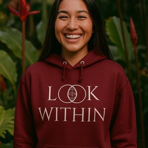 May include: A maroon hoodie with the words "LOOK WITHIN" in white, featuring a globe design within the "O"s. The hoodie has a front pocket and a drawstring hood. The person wearing the hoodie is smiling.