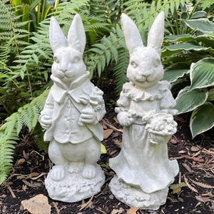 May include: Two gray stone rabbit statues. One rabbit wears a suit and holds flowers, the other wears a dress and carries a basket of flowers. Both statues have long ears and are standing on a base of leaves.