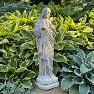 May include: A light gray stone statue of a figure with long hair and a beard, wearing draped robes. The figure stands on a rectangular base with hands positioned over the chest, featuring intricate carved details on the fabric and hair.