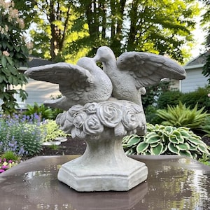 May include: A light gray stone garden statue featuring two birds with outstretched wings perched atop a cluster of sculpted roses. The birds are positioned facing each other, resting on a pedestal base with a hexagonal bottom.