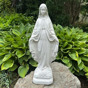 May include: A white statue of the Virgin Mary standing on a rectangular base. The figure wears a long, flowing robe and a veil covering the head, with arms extended outward at the sides. The sculpture features detailed folds in the fabric.