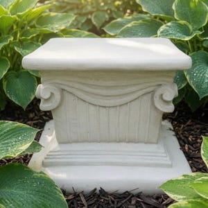 May include: A square off-white garden pedestal featuring a flat top surface, a central section with vertical fluting, and decorative scroll-like volutes on the sides. The base is wider than the top, providing a stable foundation for the structure.