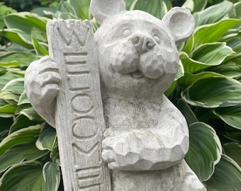 May include: A light gray stone bear statue holding a vertical rectangular sign with the word WELCOME carved into the surface. The bear has rounded ears, a textured snout, and visible carved details throughout its body, standing in a garden.