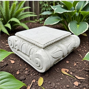 May include: A light gray stone pedestal featuring a square top with a beaded border and decorative scrollwork carvings on the sides. The base has a textured finish and is designed with symmetrical floral-inspired relief patterns on each corner.