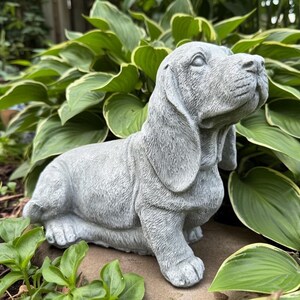 May include: A gray stone garden statue of a Basset Hound dog in a seated position. The sculpture features detailed fur texture, long drooping ears, and a wrinkled snout, capturing the characteristic facial expression of the breed in a solid form.