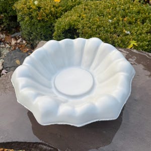 May include: A white, circular, shallow bowl with a scalloped, fluted edge and a smooth, flat center base. The object features a uniform matte finish and a distinct, wavy rim design that radiates outward from the center to the outer edge.