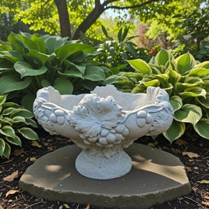 May include: A gray, stone-like planter with a pedestal base, adorned with grape and leaf relief details. The bowl-shaped top is designed for holding plants. The planter is set on a stone surface in a garden setting.