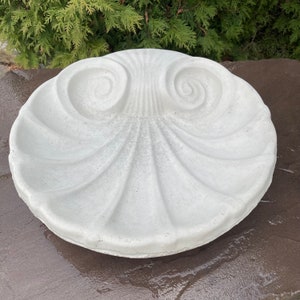 May include: A circular, off-white concrete bird bath basin featuring a molded shell design. The surface displays radiating fluted ridges that extend from a central point toward the outer edge, with two prominent spiral motifs at the top section.