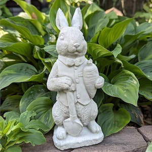 May include: A gray concrete garden statue of a rabbit wearing a vest and holding a carrot and a shovel. The rabbit has long ears and is standing on a stone base. The statue is surrounded by green foliage.