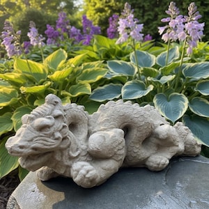 May include: A light beige stone dragon sculpture featuring a textured, scaled body, a detailed head with prominent facial features, and a curled tail. The creature is depicted in a reclining position with its limbs tucked close to its body.