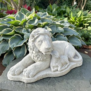 May include: A light-colored stone sculpture of a lion lying down, with a lamb resting beside it. The lion has a detailed mane and is positioned with its paws extended. The lamb is curled up next to the lion. The sculpture is placed on a large stone.