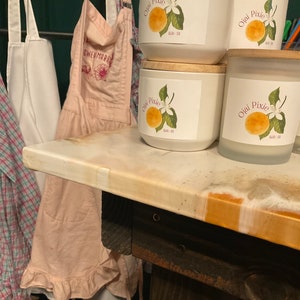 Puede incluir: Velas de cerámica blanca con tapas de madera y etiquetas que dicen "Ojai Pixie" con una ilustración de una naranja y una flor. Un delantal rosa con el texto "GWENMARK" cuelga en el fondo.