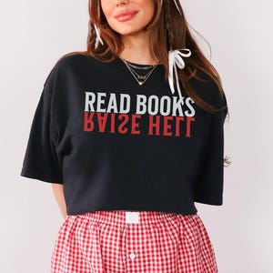 May include: A black cropped t-shirt with the text "READ BOOKS RAISE HELL" in white and red lettering. The shirt is paired with red and white gingham boxer shorts.