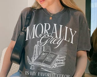 May include: Dark grey t-shirt with the words "MORALLY GREY IS MY FAVORITE COLOR" in white, along with a graphic of books and a quill. The shirt is paired with light blue jeans.