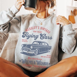 May include: Gray sweatshirt with a vintage design featuring a flying car and the text "WEASLEY'S VINTAGE Flying Cars RENT OR BUY NEW AND IMPROVED RUN OUT OF FUEL NEVER For adventurous WITCHES & WIZARDS".