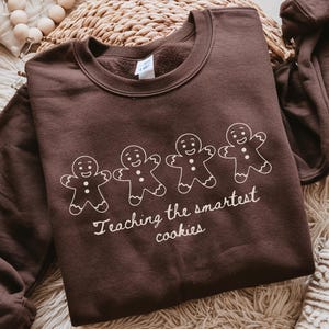 May include: A brown sweatshirt featuring a white gingerbread man design and the text "Teaching the smartest cookies." The garment is displayed on a textured surface.