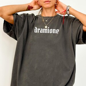 May include: Dark gray oversized t-shirt with the word "dramione" printed in white, gothic-style lettering. The shirt has short sleeves and a relaxed fit. The fabric appears to be a soft, washed material.