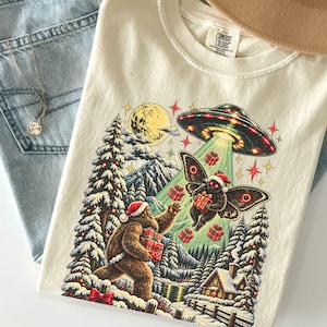 May include: An ivory t-shirt with a graphic print featuring a Bigfoot and a moth wearing Santa hats in a snowy forest. A UFO beams down red gift boxes while a full moon shines above. The shirt has a Comfort Colors label at the neck.