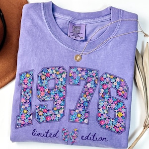 May include: Violet-colored t-shirt with the year "1976" in large, floral-filled numbers. The words "limited edition" and a floral bow are below the numbers. A gold necklace and a brown hat are also visible.