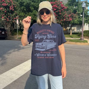 May include: A navy blue t-shirt with a vintage graphic design featuring a flying car. The graphic says "Weasley's Vintage Flying Cars" and includes the text "Rent or Buy", "New and Improved", "Never Run Out of Fuel", "For adventurous Witches & Wizards", and "The Burrow - Devon, England".