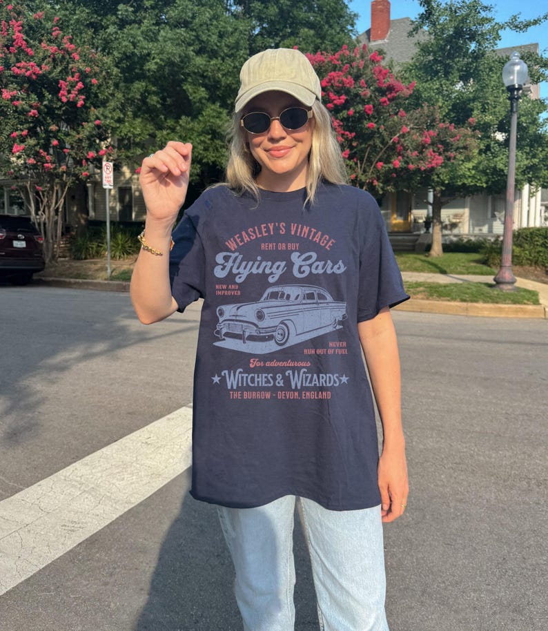 May include: A navy blue t-shirt with a vintage graphic design featuring a flying car. The graphic says "Weasley's Vintage Flying Cars" and includes the text "Rent or Buy", "New and Improved", "Never Run Out of Fuel", "For adventurous Witches & Wizards", and "The Burrow - Devon, England".